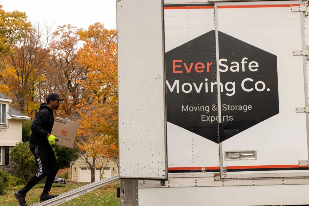 A mover in gloves walks a box up a ramp into an EverSafe Moving Co. truck surrounded by autumn leaves