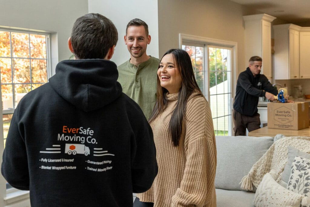 A mover in EverSafe Moving Co. gear speaks with a happy couple as another worker packs in the background