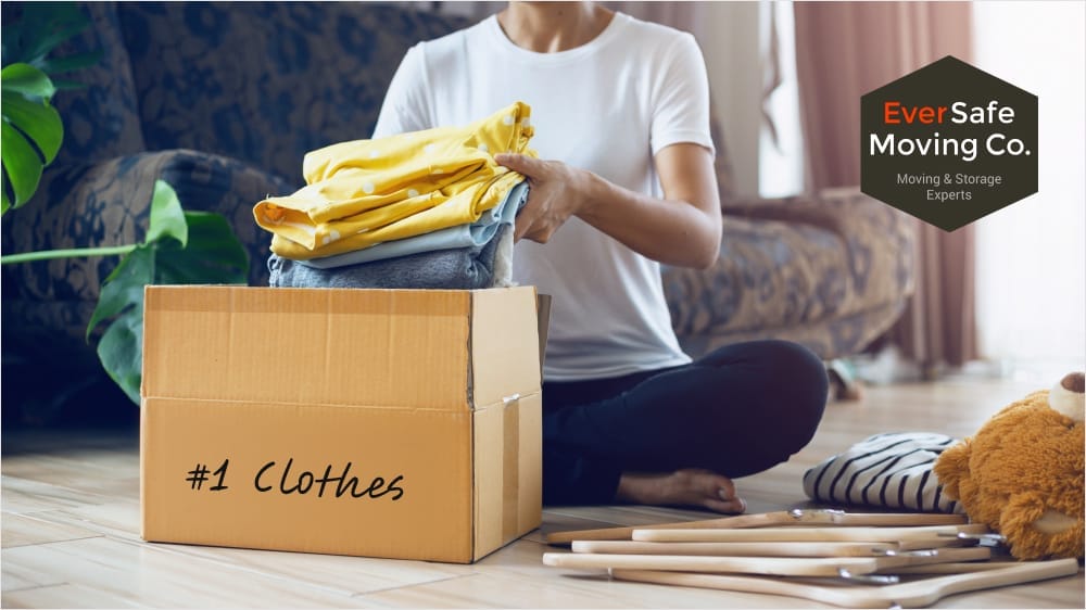 A woman placing folded clothes into a cardboard box labeled '#1 Clothes Packing,' organizing items for moving or storage.