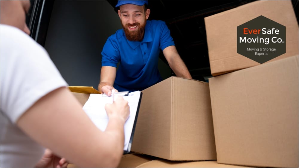 A man writing on a clipboard near the open back of a moving truck, with staff seated inside and visible packed boxes.